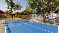 This outdoor recreational facility offers a well-maintained multi-use court surfaced in a striking blue finish with crisp white line markings, accommodating both tennis and pickleball play. The court is regulation-sized and equipped with a center net, providing a versatile space suitable for a range of racquet sports. A sturdy chain-link fence encloses the perimeter, ensuring clear boundaries and safety for players. Mature, leafy trees frame the court, offering natural shade and a pleasant, park-like atmosphere that enhances the outdoor experience. The surrounding landscape features neatly kept grassy areas, contributing to the open and inviting setting. Residential buildings constructed with classic brick and wood exteriors are visible in the backdrop, indicating the court's placement within a community environment. The open-air design takes full advantage of natural light and the surrounding greenery, making for an energizing and visually appealing environment. Overall, this facility is a well-appointed recreational amenity that serves as a focal point for outdoor activity and community engagement.