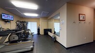 Located at 2807–2813 S Syene Rd in Fitchburg, Wisconsin, this single-floor fitness center is a well-equipped exercise facility designed to support a comprehensive workout routine. The space is finished with durable rubber flooring throughout, providing a safe and functional surface for a variety of exercises. Overhead fluorescent lighting ensures the room is consistently well-lit.

The main workout area houses a selection of cardio equipment, including a Matrix treadmill and an elliptical trainer, both positioned for convenient use and complemented by a wall-mounted television. A multi-function cable resistance machine and a dedicated weight bench round out the strength training options, while a dumbbell rack stocked with a range of free weights provides additional versatility for resistance training.

Large wall-mounted mirrors span a significant portion of one wall, visually expanding the space and allowing users to monitor their form during exercise. A water cooler station is conveniently positioned nearby, supporting hydration during workouts. Coat hooks along one wall offer practical storage, and workout reference posters are displayed throughout for exercise guidance.

The facility also includes what appears to be a separate changing or utility room, accessible via an interior door. The warm, neutral-toned walls contribute to a clean and functional atmosphere throughout the space.