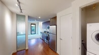 This contemporary single-floor residence offers a thoughtfully designed layout that seamlessly blends functionality with modern aesthetics. The kitchen is a standout feature, outfitted with rich walnut-toned cabinetry extending to the ceiling, dark countertops, stainless steel appliances including a refrigerator, range, microwave, and dishwasher, and warm hardwood flooring that flows throughout the unit's main areas. Recessed lighting illuminates the kitchen and living spaces, complemented by a track lighting fixture in the open living area, which transitions smoothly from hardwood to plush carpeting. The living space is bright and airy, benefiting from natural light and a view through a large black-framed window. The carpeted bedroom is generously proportioned, featuring a large window that frames lush greenery and outdoor scenery, creating a calm and serene retreat. The bathroom is clean and modern, with light tile flooring, a floating walnut vanity with an undermount sink, a framed mirror with overhead lighting, and a full toilet. Frosted glass panels near the entry add a refined architectural touch, enhancing privacy while maintaining the unit's open, contemporary feel throughout.