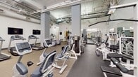This fitness center at 900-912 S Market St in Wilmington, Delaware, offers a spacious, single-floor layout designed to accommodate a comprehensive range of workout needs. The space features soaring ceilings with exposed ductwork, piping, and structural columns that lend an industrial character to the environment. Large clerestory windows allow natural light to filter in from above, brightening the predominantly white interior and contributing to an open, airy atmosphere.

The floor plan is organized to support both cardio and strength training. The cardio zone is well-stocked with treadmills, elliptical trainers, stair climbers, and recumbent bikes, all arranged in rows across carpet-tiled flooring. Wall-mounted flat-screen televisions are positioned throughout for user convenience. The strength-training area transitions to rubber flooring and houses a variety of cable machines, selectorized weight stations, and free-weight equipment, providing options for full-body conditioning.

Large mirrors are mounted along multiple walls, visually expanding the space and allowing users to monitor their form. Structural columns are integrated throughout the layout, subtly delineating workout zones. Overhead track and pendant lighting supplements natural light, ensuring consistent illumination across the entire facility. The overall ambiance is clean, functional, and professional.
