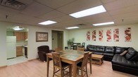 This single-floor space is designed as a multipurpose communal area, seamlessly blending a dining zone with a relaxed lounge setting. The room features a drop ceiling outfitted with recessed fluorescent panel lighting that evenly illuminates the interior, complementing the light hardwood laminate flooring that runs throughout.

The dining area is anchored by a solid wood table with a tile inlay surface, surrounded by matching wooden chairs with upholstered seats — offering both durability and comfort. Adjacent to the dining space, a cozy lounge area includes a large, dark leather sofa paired with a wooden coffee table featuring drawer storage, and a plush armchair, creating an inviting space for relaxation.

Decorative touches include a framed artwork piece on one wall and a series of four coordinating floral canvas prints on the opposite wall, adding color and visual interest. A small secondary seating area with a bistro-style table and chairs is visible in the background near a potted plant, providing an additional gathering spot. A doorway leads to a connecting room, suggesting further functional space beyond the main area.