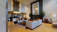 This striking contemporary lobby lounge is defined by its dramatic double-height ceilings, which lend an immediate sense of grandeur upon entry. The space is anchored by a carefully curated seating arrangement, featuring clean-lined white sofas and warm cognac accent chairs positioned around a rich wood coffee table atop a boldly patterned area rug, creating an inviting central gathering area. A large, illuminated mirror serves as a commanding focal point on the dark textured feature wall, adding depth and visual interest to the space. Lush tropical plants in deep red planters introduce vibrant color and a natural element that softens the modern aesthetic. To one side, a reception or service counter is integrated seamlessly into the design, backed by open shelving and complemented by warm wood horizontal blinds that filter natural light beautifully. Recessed lighting throughout provides a polished, even illumination, while a statement pendant fixture adds a decorative layer overhead. Light hardwood flooring runs throughout, enhancing the brightness and flow of the space. The overall ambiance is sleek and refined, balancing warmth and professionalism in equal measure.