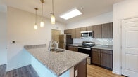 This single-story residence offers a well-designed layout with modern finishes throughout. The open-concept kitchen serves as a central feature, showcasing an expansive granite-topped island with an undermount sink, dark wood-tone cabinetry, and a full suite of stainless steel appliances including a refrigerator, range, and over-the-range microwave. A white subway tile backsplash adds a clean contrast to the cabinetry, while pendant lighting and a recessed ceiling light illuminate the workspace effectively. Wood-look plank flooring extends through the kitchen and living areas, lending warmth and continuity. The living room is generously proportioned, featuring a ceiling fan, neutral walls, and windows that bring in natural light with views of the surrounding area. A private exterior door provides direct outdoor access. Bedrooms are finished with plush carpeting, ceiling fans, and windows offering natural light. The bathrooms mirror the kitchen's design aesthetic, featuring matching granite vanity countertops, dark cabinetry with ample storage, wood-look flooring, and a shower-tub combination. A dedicated laundry area is also visible within the unit, adding practical convenience to the overall layout.