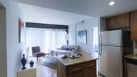 This single-floor residence showcases an open-concept layout that connects the kitchen and living areas in one cohesive space. The kitchen features a substantial island topped with granite countertops and rich dark wood cabinetry with brushed metal hardware, complemented by a stainless steel refrigerator. The design flows naturally into the living area, which is bathed in abundant natural light streaming through expansive floor-to-ceiling windows dressed with sheer vertical blinds. The living space is generously proportioned, offering room for a full seating arrangement including a sofa accented with purple throw pillows and a coordinating accent chair. Light hardwood flooring runs throughout, adding warmth to the neutral white walls and creating a clean, contemporary aesthetic. A framed artwork piece anchors one wall of the living area, while a glass-top coffee table and area rug define the seating zone. The open layout and bright, airy atmosphere make this a well-connected and functional living environment suited for both everyday living and entertaining.