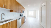 This single-floor unit showcases a contemporary design aesthetic characterized by clean lines, a neutral palette, and warm natural wood tones throughout. The galley-style kitchen is a standout feature, lined with rich wood cabinetry extending from floor to ceiling and fitted with brushed gold hardware. Stainless steel appliances — including a range, over-the-range microwave, dishwasher, and refrigerator — provide a polished, modern finish, while a white quartz countertop and a distinctive scalloped white tile backsplash add texture and visual interest. Light hardwood flooring flows continuously from the kitchen into the open living area, reinforcing a cohesive, airy feel.

The living space is bathed in natural light, drawing the eye toward a set of large windows that frame expansive urban skyline views — a particularly notable feature of the unit. Recessed lighting throughout ensures consistent illumination across all areas.

The bathroom is finished in a clean, minimalist style with full subway tile surrounding the soaking tub and shower, a floating vanity with a gray wood-tone base, a wide white basin sink, and a large mirror. The overall ambiance of the unit is bright, modern, and thoughtfully designed, offering a well-balanced blend of function and contemporary style.