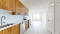 This single-floor unit showcases a contemporary design aesthetic characterized by clean lines, a neutral palette, and warm natural wood tones throughout. The galley-style kitchen is a standout feature, lined with rich wood cabinetry extending from floor to ceiling and fitted with brushed gold hardware. Stainless steel appliances — including a range, over-the-range microwave, dishwasher, and refrigerator — provide a polished, modern finish, while a white quartz countertop and a distinctive scalloped white tile backsplash add texture and visual interest. Light hardwood flooring flows continuously from the kitchen into the open living area, reinforcing a cohesive, airy feel.

The living space is bathed in natural light, drawing the eye toward a set of large windows that frame expansive urban skyline views — a particularly notable feature of the unit. Recessed lighting throughout ensures consistent illumination across all areas.

The bathroom is finished in a clean, minimalist style with full subway tile surrounding the soaking tub and shower, a floating vanity with a gray wood-tone base, a wide white basin sink, and a large mirror. The overall ambiance of the unit is bright, modern, and thoughtfully designed, offering a well-balanced blend of function and contemporary style.