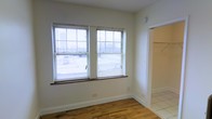 This single-floor space presents a straightforward and functional layout with a clean, neutral aesthetic throughout. The main room features warm hardwood flooring that adds character to the otherwise minimalist design, with crisp white walls that keep the space feeling open and bright. Two side-by-side windows fitted with horizontal blinds allow generous natural light to fill the room, providing views of the surrounding exterior environment. The white baseboard trim provides a classic finishing detail along the perimeter of the room. Adjacent to the main area, a transition to tile flooring marks the entry into a secondary space, which includes an open closet area with visible hanging storage — practical and easily accessible. The overall ambiance is simple and uncluttered, making the space highly adaptable for a variety of uses. The combination of hardwood and tile flooring across areas adds both warmth and practicality, while the abundance of natural light ensures the interior feels airy and welcoming throughout the day.