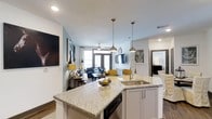 This two-bedroom, two-bathroom residence offers a thoughtfully designed single-floor layout that maximizes functionality and flow. The open-concept design connects the kitchen, dining, and living areas into one cohesive, well-lit space, creating an inviting atmosphere throughout.

The kitchen serves as a central hub, anchored by a large granite-topped island with an undermount sink, pendant lighting with gold-toned fixtures, and white shaker-style cabinetry. A dishwasher is integrated beneath the island, and the space transitions seamlessly into the dining and living areas.

The living area benefits from abundant natural light streaming through large windows and French doors, which lead to an outdoor access point. A ceiling fan adds comfort, while the open sightlines enhance the sense of spaciousness. The light blue-gray wall tones complement the warm wood-style flooring found throughout the residence.

Overall, the space balances modern design elements — including recessed lighting, contemporary fixtures, and clean architectural lines — with a warm, livable ambiance suited for everyday comfort.