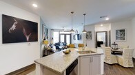 This two-bedroom, two-bathroom residence offers a thoughtfully designed single-floor layout that maximizes functionality and flow. The open-concept design connects the kitchen, dining, and living areas into one cohesive, well-lit space, creating an inviting atmosphere throughout.

The kitchen serves as a central hub, anchored by a large granite-topped island with an undermount sink, pendant lighting with gold-toned fixtures, and white shaker-style cabinetry. A dishwasher is integrated beneath the island, and the space transitions seamlessly into the dining and living areas.

The living area benefits from abundant natural light streaming through large windows and French doors, which lead to an outdoor access point. A ceiling fan adds comfort, while the open sightlines enhance the sense of spaciousness. The light blue-gray wall tones complement the warm wood-style flooring found throughout the residence.

Overall, the space balances modern design elements — including recessed lighting, contemporary fixtures, and clean architectural lines — with a warm, livable ambiance suited for everyday comfort.