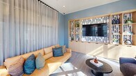 This 190 square foot living room exemplifies modern urban design with its efficient use of space and contemporary aesthetics. The room is bathed in natural light thanks to large windows that offer views of the surrounding area. The walls are painted in a soothing blue-gray tone, creating a calm and sophisticated ambiance.

The space features a versatile seating arrangement, including a vibrant yellow sofa adorned with blue and gray pillows, and two comfortable brown armchairs. A round coffee table centers the seating area, while a floor lamp adds focused lighting. The room's flooring consists of a neutral-toned carpet with a subtle pattern, adding texture without overwhelming the space.

A standout feature is the floor-to-ceiling sheer curtain that can be drawn to divide the room or provide privacy. The room also includes built-in shelving and a media center, cleverly integrating storage and entertainment options. Recessed lighting in the ceiling provides even illumination throughout the space.

The entrance area features a glass door, allowing for additional natural light and a seamless transition to the exterior. Overall, this living room demonstrates a perfect balance of style, comfort, and functionality in a compact urban setting.