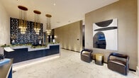 This elegantly designed lobby and hallway serves as the welcoming centerpiece of a modern residential building. The space is grounded by expansive white Calacatta marble-style flooring, whose soft veining lends a refined, upscale character from entry to elevator corridor. At the heart of the lobby stands a sleek reception desk finished in navy fluted paneling with a light marble counter, accented by cascading brass pendant chandeliers that add warmth and drama overhead. The surrounding walls are clad in rich taupe textured wallcovering, complemented by a bold three-dimensional geometric black tile feature wall adorned with decorative vases — a striking focal point that adds depth and artistry to the space. A seating area furnished with curved leather armchairs and a brushed gold accent table provides a comfortable waiting area, positioned near a modern abstract wall panel and an integrated digital display. The hallway extends toward two sleek elevator bays, flanked by wall sconces and additional digital screens for resident convenience. Near the main entry, glass double doors flood the space with natural light, while a console table and mounted display screen offer an informative welcome. The overall ambiance is polished, contemporary, and inviting — reflecting a high standard of design throughout.