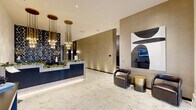 This elegantly designed lobby and hallway serves as the welcoming centerpiece of a modern residential building. The space is grounded by expansive white Calacatta marble-style flooring, whose soft veining lends a refined, upscale character from entry to elevator corridor. At the heart of the lobby stands a sleek reception desk finished in navy fluted paneling with a light marble counter, accented by cascading brass pendant chandeliers that add warmth and drama overhead. The surrounding walls are clad in rich taupe textured wallcovering, complemented by a bold three-dimensional geometric black tile feature wall adorned with decorative vases — a striking focal point that adds depth and artistry to the space. A seating area furnished with curved leather armchairs and a brushed gold accent table provides a comfortable waiting area, positioned near a modern abstract wall panel and an integrated digital display. The hallway extends toward two sleek elevator bays, flanked by wall sconces and additional digital screens for resident convenience. Near the main entry, glass double doors flood the space with natural light, while a console table and mounted display screen offer an informative welcome. The overall ambiance is polished, contemporary, and inviting — reflecting a high standard of design throughout.