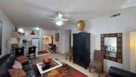 This single-floor residence offers a thoughtfully arranged open-concept layout that connects the living area, dining space, and kitchen in one cohesive flow. The living room serves as the centerpiece of the home, featuring light-colored carpeting and a ceiling fan with integrated lighting that enhances both comfort and ambiance. The space is furnished with a mix of bold, eclectic pieces, including a striking dark armoire, a glass-top coffee table, and richly patterned area rugs in warm red and brown tones that add depth and character to the room. A cozy seating arrangement anchors the living area, creating an inviting atmosphere for relaxing or entertaining. Just beyond, the dining area provides an intimate setting with a compact table and chairs, accented by decorative wall art. The kitchen is visible in the background, featuring white appliances and an efficient layout. A built-in desk or workstation area is also visible, offering a dedicated functional nook within the open floor plan. Throughout the space, white walls and consistent flooring create a clean, unified backdrop that allows the warm furnishings and accessories to stand out. The overall ambiance is cozy and lived-in, with a welcoming character defined by its rich color palette and layered décor.
