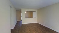 This single-floor residence offers a clean, functional layout with a welcoming sense of openness throughout. The main living area is anchored by rich, dark hardwood-style flooring that runs continuously underfoot, providing warmth and visual depth. Neutral, light-toned walls keep the space feeling bright and versatile, ready to complement a variety of interior styles.

A standout architectural detail is the built-in pass-through window cut into the interior wall, which visually connects the living area to an adjacent room. This design element enhances the open feel of the floor plan and allows light and sightlines to flow freely between spaces. Through the pass-through, a ceiling fan is visible, hinting at the well-appointed character of the connected room.

A hallway extends from the living area toward a naturally lit entryway, where vertical blinds soften incoming daylight. The textured ceiling adds a subtle layer of dimension, and clean white baseboards frame the flooring throughout. The overall ambiance is simple, well-maintained, and move-in ready, offering a neutral canvas suited to a range of personal styles and furnishing preferences.