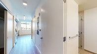 This single-floor unit combines modern finishes with a functional layout across several well-defined areas. The corridor serves as the central spine of the space, featuring white walls, light wood-style flooring, and multiple doors with stainless steel hardware. Notably, the hallway includes a keypad-secured entry door, adding a layer of access control, and is illuminated by recessed linear lighting that enhances its clean, contemporary feel.

The kitchen is a standout area, outfitted with rich dark-stained cabinetry extending to the ceiling, light granite countertops, and a white mosaic tile backsplash. A full suite of stainless steel appliances—including a gas range, built-in microwave, refrigerator, and dishwasher—provides modern functionality. The flooring here continues the light wood-style planks seen in the corridor.

The bedroom features neutral carpet, a large window that invites natural light and exterior views, and an open closet with a hanging rod. The room's simple, bright design creates a calm and versatile space.

The bathroom is finished with large-format tile flooring, a dark-cabineted vanity topped with granite, a wide mirror, and a glass-enclosed shower with accent tile detailing. Grab bars are thoughtfully incorporated, indicating accessibility-conscious design throughout the unit.