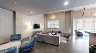 Located on Holmes Ave NW in Huntsville, Alabama, this single-floor space masterfully combines industrial character with contemporary elegance. Soaring ceilings are supported by exposed concrete columns, while polished concrete floors run throughout, creating a cohesive, loft-inspired aesthetic. A dramatic crystal chandelier anchors the main lounge area, where generous seating arrangements—including plush sofas, accent chairs upholstered in bold blue patterned fabric, and modern armchairs—invite relaxed gathering. Large windows fitted with sheer roller shades bathe the interior in abundant natural light while offering views of the tree-lined streetscape outside.

The bar area serves as a striking focal point, featuring a broad concrete countertop edged with a herringbone wood-paneled base, white upholstered swivel barstools, and a full mirrored subway tile backsplash reflecting pendant lighting overhead. Dual wall-mounted televisions provide visual entertainment throughout. Behind the bar, rich dark wood cabinetry with brushed steel hardware and additional concrete countertops offer substantial functional workspace. Industrial-style open shelving adds a curated, editorial touch. The overall ambiance is polished yet approachable, suited for events, social gatherings, or professional functions.