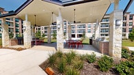 Located at 15915 Kingfield Dr in Houston, TX, this expansive outdoor amenity area offers a well-designed communal environment set against the backdrop of a modern multi-story residential building. The centerpiece of the space is a generously sized covered pavilion supported by natural stone columns and topped with a standing-seam metal roof, providing shade and shelter for outdoor gatherings. Ceiling fans and pendant lighting ensure comfort throughout the day and into the evening.

Beneath the pavilion, multiple dining tables and vibrant red chairs create inviting social areas, while a built-in outdoor grilling station with adjacent counter space makes al fresco entertaining convenient. Curved concrete walkways wind through the grounds, connecting the pavilion to additional outdoor features including a putting green with artificial turf, a children's playground with a slide, and raised garden or seating areas with bold red benches.

The grounds are thoughtfully landscaped with manicured lawns, ornamental grasses, native shrubs, and young trees that add greenery and seasonal interest. An interior lounge area complements the outdoor amenities, featuring white walls accented by bold artwork, a central round table with red seating, and a clean, open layout. Together, these spaces create a vibrant and functional community amenity destination.
