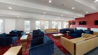 This single-floor community amenity space is thoughtfully designed to serve as a vibrant social hub. The layout is divided into two distinct yet complementary areas, each offering its own character and function.

The lounge area is expansive and airy, with vaulted ceilings that amplify the sense of openness. A bold red accent wall serves as a striking focal point, complemented by two sleek wall-mounted flat-screen TVs. The seating arrangement is both stylish and inviting, featuring a tufted caramel leather sofa, rich navy blue velvet armchairs and sofas, and gold-toned accent pieces, all anchored by vibrant patterned area rugs. Dramatic sputnik-style chandeliers hang overhead, adding a mid-century modern flair. Floor-to-ceiling windows along one wall flood the space with natural light and offer clear views of an outdoor pool area, accessible through a set of double glass doors.

The kitchenette and bar area transitions seamlessly from the lounge and is finished with white shiplap walls, light hardwood-style flooring, and crisp white cabinetry. Vivid red countertops on both the bar island and the back counter create a cohesive, energetic color story. A pendant light in matching red anchors the island, while a row of wall-mounted TVs above the counter makes this area ideal for entertaining and watching events.