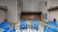 Located at 30 S Court St in Athens, Ohio, this single-floor property offers a notable outdoor living space that serves as a central highlight. The exterior features a generous wooden deck area nestled within the building's courtyard-style layout, creating a semi-enclosed outdoor environment that provides a sense of privacy and community. The deck is outfitted with a covered section supported by wooden posts and framed with decorative lattice railings, offering shade and shelter for gatherings. Adirondack-style seating in bold blue adds a welcoming, casual atmosphere to the space, complemented by a striped outdoor rug that defines the seating area. The surrounding structure is clad in neutral-toned horizontal siding, with multiple windows overlooking the deck from multiple sides, allowing natural light to flow into the adjacent interior spaces. The multi-wing building design creates an intimate courtyard feel, making the outdoor deck a natural gathering hub. The overall ambiance is relaxed and functional, well-suited for casual outdoor enjoyment in the heart of Athens, Ohio.