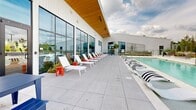 Located at 1200 Foster St NW in Atlanta's dynamic West Midtown neighborhood, this rooftop pool deck offers a compelling blend of modern design and resort-style amenities. The centerpiece is a generously sized swimming pool framed by an expansive concrete deck, lined with white lounge chairs and boldly accented side tables, creating a vibrant and inviting atmosphere for relaxation.

The building's exterior showcases crisp white corrugated metal cladding paired with large floor-to-ceiling windows and a warm wood-paneled ceiling overhang, lending the space a contemporary industrial aesthetic. Integrated planters and lush greenery — including ornamental grasses, flowering plants, and upright evergreen trees — soften the hardscape and contribute to a rooftop garden feel.

Seating areas are thoughtfully distributed throughout the deck, from sleek slatted wood benches nestled among the plantings to lounge groupings with modern wire chairs and colorful accent tables. A separate patio area offers a blue picnic table set against a backdrop of open sky and greenery.

The rooftop commands impressive panoramic views of the Atlanta skyline, with the city's distinctive skyline clearly visible across the horizon. Glass railings preserve sightlines while maintaining an open, airy ambiance. This space is ideal for outdoor leisure, social gatherings, and soaking in Atlanta's dynamic urban landscape.