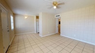 This single-story space features an open, airy layout finished with light beige ceramic tile flooring throughout, lending a clean and cohesive aesthetic. The main living area is generously proportioned, offering flexibility in furniture arrangement and everyday use. Distinctive textured block walls line the perimeter, adding architectural character and a subtle mid-century feel to the interior. A ceiling fan with a brass-toned fixture is centrally mounted on the textured white ceiling, providing both airflow and ambient lighting to the space. A central dividing wall partially separates the living area from the kitchen zone, which is visible in the background and includes a white refrigerator tucked neatly into a recessed alcove. A wall-mounted light fixture adds supplemental lighting near the kitchen transition. A white interior door leads to additional rooms beyond, suggesting a practical and connected floor plan. Overhead HVAC venting is integrated into the ceiling, supporting climate control throughout. The overall ambiance is bright, neutral, and functional, providing a versatile canvas suitable for a variety of uses.