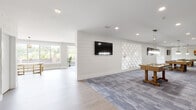 This single-floor amenity space is thoughtfully designed to accommodate a variety of social and recreational activities across several well-defined zones. The overall aesthetic is clean and contemporary, characterized by light hardwood-style flooring, soft neutral wall tones, and recessed lighting throughout.

The lounge area is bathed in natural light from a generous bank of large windows overlooking an outdoor terrace with landscaped plantings. Seating here includes a teal sofa accented with green and grey pillows, paired with mid-century-style armchairs and a marble-topped accent table with a gold pedestal base, creating an inviting and upscale atmosphere.

A dedicated dining nook features botanical-patterned wallpaper, pendant lighting, a marble bistro table, and gold wire-frame chairs beneath a vibrant abstract art piece, adding an artistic focal point to the space.

The expansive game room is anchored by a shiplap-clad accent wall with a mounted flat-screen TV, a diamond-pattern textured wall panel, and grey patterned carpet tiles. Foosball and billiards tables are centrally positioned, while gold bar-height stools line the perimeter for spectating. Together, these areas form a cohesive, amenity-rich environment ideal for socializing and recreation.