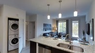 This single-floor space showcases a thoughtfully designed open-concept layout that seamlessly connects the kitchen and living areas. The kitchen is a standout feature, centered around a large island with light granite countertops, a deep undermount double sink with a sleek pull-down faucet, and dark cabinetry that provides a striking contrast to the bright, neutral walls. A stainless steel dishwasher is integrated into the island base, adding to the kitchen's modern functionality. Three pendant lights with cylindrical shades hang above the island, casting warm, focused illumination over the workspace and dining surface.

The open layout flows naturally into the living area, which is anchored by large windows and a glass door that flood the space with natural light and offer views of the surrounding greenery. The living area features a comfortable leather seating arrangement, complemented by tasteful décor including a geometric wall art piece and ambient floor and table lamps. Light wood-style flooring runs throughout, tying the spaces together cohesively. A hallway visible from the kitchen suggests additional rooms beyond, completing this well-appointed and inviting single-floor residence.