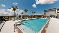 Located at 606 SW 29th Ave, this single-floor community amenity space offers a well-appointed interior complemented by an expansive outdoor pool area. The interior showcases a clean, coastal aesthetic with light wood-look flooring, crisp white walls, and an open layout that seamlessly combines the living and dining areas. The living space is furnished with contemporary teak-framed lounge chairs dressed in white cushions and navy accents, centered around ocean-inspired artwork that reinforces the coastal theme. A coffee bar area provides a functional refreshment station complete with counter seating, dual mounted televisions set against a wood-paneled accent wall, and under-counter refrigeration. The dining area features a round white table bathed in natural light from large windows adorned with plantation shutters, with vivid blue wave-motif artwork adorning the walls. Glass-paneled interior doors connect the various zones, maintaining an open and airy feel throughout. Outside, the resort-style pool area is a standout feature, offering a large sparkling pool and a separate spa, both surrounded by a spacious deck lined with teal and white lounge chairs. Towering palm trees, an accessible pool lift, and iron perimeter fencing complete the inviting outdoor retreat, all set beneath the open Florida sky.