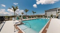 Located at 606 SW 29th Ave, this single-floor community amenity space offers a well-appointed interior complemented by an expansive outdoor pool area. The interior showcases a clean, coastal aesthetic with light wood-look flooring, crisp white walls, and an open layout that seamlessly combines the living and dining areas. The living space is furnished with contemporary teak-framed lounge chairs dressed in white cushions and navy accents, centered around ocean-inspired artwork that reinforces the coastal theme. A coffee bar area provides a functional refreshment station complete with counter seating, dual mounted televisions set against a wood-paneled accent wall, and under-counter refrigeration. The dining area features a round white table bathed in natural light from large windows adorned with plantation shutters, with vivid blue wave-motif artwork adorning the walls. Glass-paneled interior doors connect the various zones, maintaining an open and airy feel throughout. Outside, the resort-style pool area is a standout feature, offering a large sparkling pool and a separate spa, both surrounded by a spacious deck lined with teal and white lounge chairs. Towering palm trees, an accessible pool lift, and iron perimeter fencing complete the inviting outdoor retreat, all set beneath the open Florida sky.