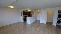 This single-floor space offers a thoughtfully designed open-concept layout that maximizes functionality and flow. The living area is expansive and airy, featuring light wood-style luxury vinyl flooring that runs throughout, creating a cohesive and modern aesthetic. Crisp white walls enhance the brightness of the space, reflecting light to create a welcoming ambiance.

The kitchen is positioned at the heart of the unit, defined by a white peninsula counter that doubles as a breakfast bar and provides a natural division between the kitchen and living areas. Dark cabinetry offers a striking contrast against the white walls and countertops, adding depth and a contemporary edge to the design. The kitchen is equipped with stainless steel appliances, including a refrigerator and microwave, along with a black range, providing a complete and functional cooking space.

A utility or electrical panel is discreetly integrated into the wall near the kitchen, maintaining the clean lines of the interior. An entryway door with a deadbolt is visible toward the rear of the space, offering a defined entry and exit point. Flush-mount ceiling light fixtures provide consistent, even illumination throughout. Overall, the space strikes a balance between modern style and practical livability.