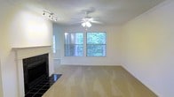 This single-floor space presents a welcoming living area designed with both comfort and functionality in mind. The main living space is finished with wall-to-wall carpeting in a neutral tone, creating a warm and inviting atmosphere. A standout feature is the classic fireplace with a dark tile surround and a clean white mantle, serving as a natural focal point within the room. Adjacent to the fireplace, a set of track lights mounted on the wall provides additional directional lighting, complementing the ceiling fan with an integrated light fixture that occupies the center of the room. Two generously sized windows allow abundant natural light to fill the space and offer pleasant views of the surrounding greenery, giving the interior a bright, airy feel. The textured ceiling adds a traditional architectural detail consistent throughout the space. The neutral white walls provide a versatile backdrop, suitable for a variety of décor styles. Overall, the space carries a clean, functional character with practical features well-suited for everyday living.