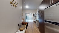 This single-floor studio unit offers a well-organized, open-concept layout that efficiently combines living, sleeping, and cooking areas within one cohesive space. Light-toned hardwood-style flooring runs continuously throughout, contributing to a clean and cohesive aesthetic.

The main living area is bright and airy, anchored by a large sliding glass door that opens onto a private balcony, allowing natural light to fill the room and providing an outdoor retreat. The sleeping area features a rattan daybed that doubles as a seating space, complemented by framed wall art, adding a relaxed, coastal ambiance.

The entryway is functional and welcoming, with a wall-mounted coat rack and a storage bench, providing practical organization from the moment of arrival. Adjacent to the entry, the kitchen is compact yet fully equipped, featuring dark espresso-toned cabinetry that reaches to the ceiling, granite countertops, a stainless steel refrigerator, microwave, and a sink — offering all essential cooking amenities.

The bathroom includes a full-sized tub and shower combination with clean white surround panels, chrome fixtures, and wood-style flooring that ties in with the rest of the unit. Overall, the space presents a modern, well-appointed design that balances functionality with a warm, inviting atmosphere.