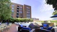 Located at 415-425 N Front St in Columbus, Ohio, this outdoor amenity area is situated at the base of a multi-story brick building in the heart of the Short North/Downtown Columbus corridor. The space is designed to serve as a communal gathering and relaxation destination, combining multiple functional zones within a well-landscaped courtyard setting.

At the center of the space is a rectangular swimming pool surrounded by a broad pool deck furnished with rows of lounge chairs, ideal for sunbathing and leisure. Adjacent to the pool area is a dedicated lounge zone featuring wicker sectional seating with bold blue cushions, arranged around a domed fire pit that serves as a striking focal point for evening gatherings.

Lush greenery and mature shrubs line the perimeter, providing a natural buffer that adds privacy and softens the urban surroundings. The brick facade of the building rises prominently overhead, with large windows and private balconies visible on upper floors. The overall ambiance balances an active outdoor lifestyle with comfortable, resort-inspired relaxation in an urban Columbus setting.