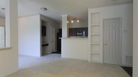 This single-floor residence offers a thoughtfully designed open-concept layout that seamlessly connects the living and kitchen areas. The living space features plush wall-to-wall carpeting and crisp white walls, creating a clean and welcoming atmosphere. A defining architectural feature is the decorative white column with crown molding detail that frames the transition into the kitchen area, adding a touch of elegance to the open floor plan. Built-in shelving units along one wall provide practical display and storage space, while a nearby door offers convenient access to an additional room or closet. The kitchen is anchored by a raised peninsula with a light granite-style countertop, ideal for casual dining or food preparation. Dark cabinetry provides a striking contrast against the lighter walls, and the space is equipped with modern appliances including a microwave and refrigerator. Track lighting above the kitchen ensures ample illumination. Beyond the kitchen, a hallway leads to additional rooms, with glimpses of more cabinetry and a built-in desk or counter area visible in the background. The overall ambiance is bright, functional, and move-in ready.