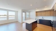 Unit 306 at 3099 Iowa Ave is a thoughtfully designed 590-square-foot, one-bedroom, one-bathroom apartment located on the third floor in Riverside, CA. The open-concept layout is anchored by a modern kitchen featuring warm wood upper cabinetry, two-tone lower cabinets in a bold navy-blue finish, a quartz waterfall island, stainless steel appliances including a French door refrigerator, electric range, built-in microwave, and dishwasher, plus a subway tile backsplash and matte black fixtures. Light wood-tone flooring flows seamlessly throughout the entire unit, enhancing the bright, airy aesthetic. The 120-square-foot primary bedroom is bathed in natural light through large sliding windows that offer outdoor views, and includes a ceiling fan and access to a dedicated closet. The bathroom is finished with a floating wood vanity, a large mirror with overhead lighting, open shelving, and a glass-enclosed walk-in shower. Recessed lighting, clean white walls, and a well-proportioned entrance complete the unit. The property also includes an assigned covered parking space, visible in the aerial overview of the building.