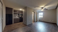 Located at 2616 N Kansas Expy in Springfield, Missouri, this single-floor unit offers a thoughtfully designed open-concept layout that connects the kitchen and living areas seamlessly. Dark hardwood-style flooring extends throughout the space, providing a cohesive and modern aesthetic.

The kitchen is equipped with black appliances, including a range, microwave, and dishwasher, complemented by dark wood cabinetry with sleek hardware and an open display shelf. A light-toned countertop provides contrast against the darker elements, and a textured accent wall with tile-style backsplash adds visual depth to the space.

The living area is bright and open, featuring a ceiling fan with a light fixture and a wall-mounted HVAC unit. A window allows natural light to filter in, enhancing the airy feel of the room. A partially open doorway provides access to additional areas of the unit, including a dedicated laundry closet.

The bedroom is a generously proportioned, quiet space with the same dark hardwood-style flooring, neutral gray walls, and exposed beam detailing near the ceiling that adds a subtle architectural character.

The bathroom features coordinating dark wood cabinetry, a vanity with a white countertop and sink, a large mirror, wall-mounted storage cabinet, and hardwood-style flooring that maintains the design continuity found throughout the rest of the unit.