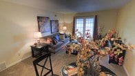 This single-floor living space offers a cozy and well-appointed interior designed for comfortable everyday living. The main living area is carpeted and centers around a gray sofa accented with colorful throw pillows, complemented by a dark wood side table and warm ambient lighting from table lamps. Two large, vibrant abstract paintings anchor the wall, adding a bold artistic touch to the neutral color palette of beige walls and dark trim. A set of French doors at the far end of the room invites natural light deep into the space while offering an outdoor view, framed by rich brown curtain panels that add warmth and dimension. The foreground reveals a dining area with dark wood seating and a decorative table arrangement featuring autumn-toned floral centerpieces, reinforcing the warm, seasonal ambiance throughout. The open layout between the dining and living areas creates a sense of flow and spaciousness, making the space feel welcoming and functional. Overall, the interior balances decorative flair with practical furnishings, resulting in a homey atmosphere with thoughtful attention to detail in its styling.