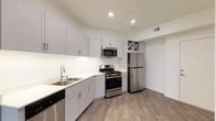 This well-appointed single-floor residence combines contemporary design with functional living. The kitchen is a standout feature, outfitted with crisp white shaker-style cabinetry that extends to the ceiling, providing generous storage throughout. Stainless steel appliances — including a gas range, over-the-range microwave, refrigerator, and dishwasher — lend a polished, modern aesthetic. The white quartz countertops and double-basin sink offer ample workspace, while recessed lighting illuminates the room with a clean, bright ambiance. A built-in wine rack adds a thoughtful decorative touch above the refrigerator. Warm wood-look flooring runs throughout, grounding the space with a welcoming feel that complements the neutral color palette. The private balcony extends the living experience outdoors, framed by a classic arched stucco surround. From here, residents enjoy elevated views of swaying palm trees, a lushly landscaped courtyard, and the surrounding community buildings set against an open sky with mountain silhouettes visible in the distance. The overall design balances modern finishes with a comfortable, resort-inspired atmosphere ideal for everyday living.