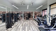 This fitness center offers a generously sized, open-concept layout designed to accommodate a full range of workout activities. The space features durable wood-look plank flooring in neutral tones, lending a clean and modern aesthetic throughout. Overhead, multiple ceiling fans complement recessed and linear LED lighting, ensuring the room remains well-lit and comfortable during use.

The facility is organized to maximize functionality, with cardio equipment — including a row of treadmills — positioned along one side, while strength training machines and multi-use cable systems are distributed across the central and opposite areas of the room. Structural columns are integrated seamlessly into the layout without significantly impeding the open feel of the space.

Large windows along one wall allow natural light to filter into the room, contributing to a bright and energizing atmosphere. Wall-mounted mirrors enhance the sense of space and support proper form during exercise. The neutral color palette of soft grays and whites creates a clean, professional environment. Overall, this fitness center is thoughtfully arranged to support a variety of training needs in a comfortable, modern setting.