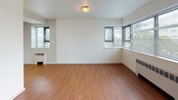 This single-floor residence offers a straightforward, functional layout with a clean, neutral aesthetic throughout. The living area is generously proportioned, featuring warm-toned hardwood-style flooring and crisp white walls that create a bright, open atmosphere. Two windows fitted with horizontal blinds allow natural light to fill the space while offering views of surrounding greenery.

The galley-style kitchen runs along two parallel walls, maximizing storage and workspace efficiency. White cabinetry with gold-tone hardware lines both sides from floor to ceiling, complemented by white countertops, a built-in electric range, and a single-basin sink. Overhead fluorescent lighting ensures the space is well-lit throughout.

The bedroom is finished with neutral wall-to-wall carpeting and features a window with blinds, providing both comfort and natural light. A wall-mounted heating unit is positioned beneath the window, offering practical climate control.

The bathroom is cleanly appointed with hexagonal tile flooring, a single-sink vanity with under-cabinet storage, a large mirror with overhead bar lighting, a full-size soaking tub with a tiled surround, and standard fixtures throughout. The overall design of the unit prioritizes functionality, with consistent white walls and light flooring unifying the space.