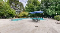 Located at Club Lakes in Lawrenceville, Georgia, this outdoor pool amenity area offers a well-appointed retreat surrounded by mature trees and dense natural greenery. The centerpiece is a generously sized swimming pool with clear turquoise water, accessible via handrail-equipped steps and framed by a broad concrete pool deck. The deck provides ample space for lounging, with multiple sling-style chaise lounge chairs positioned to take in the serene, wooded backdrop. A dedicated umbrella and seating area offers additional shade for relaxing outdoors. The entire pool area is enclosed by a sturdy black iron fence with a gated entry, ensuring both security and an aesthetic boundary between the pool and surrounding landscape. Adjacent to the pool, a single-story blue-gray utility building houses what appear to be restroom and mechanical facilities, finished with white doors and covered by an overhead overhang. Lush shrubs and ornamental plantings line the perimeter, softening the transition between the built environment and the natural tree canopy overhead. Towering pines and hardwoods create a private, park-like atmosphere that defines the overall ambiance of the space, making it a tranquil outdoor destination for residents of the Club Lakes community.