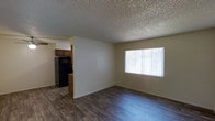 This single-floor residence offers a straightforward and functional layout designed for comfortable everyday living. The open living area is finished with durable wood-look laminate flooring in a rich, dark tone that runs throughout the space, adding warmth and visual continuity. A large window fitted with vertical blinds allows natural light to fill the room, creating a bright and inviting atmosphere. The textured popcorn ceiling adds a classic touch overhead. Adjacent to the living area, the kitchen is partially visible and features warm wood-toned cabinetry, a dark refrigerator, and a matching dark range, providing a practical cooking space. The kitchen is separated from the living area by a partial wall with a countertop pass-through, maintaining an open feel while defining each area. The neutral, light-toned walls throughout the space offer a clean and versatile backdrop, ready to accommodate a variety of interior styles. The overall layout is efficient and well-organized, making thoughtful use of the available space on a single level.