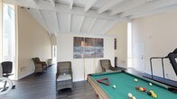 This single-story space is designed as a dedicated recreational and entertainment area, combining casual comfort with functional design. The room's most notable architectural feature is its vaulted ceiling with white-painted exposed wood beams, which adds visual height and an open, airy character to the interior. Dark hardwood flooring grounds the space and provides a rich contrast to the bright, neutral walls and ceiling.

At the center of the room, a full-sized billiard table with a green felt surface serves as the focal point, accompanied by cues and billiard accessories ready for use. Woven rattan lounge chairs are positioned around the room, offering comfortable seating for guests. A striking triptych wall art piece depicting a dramatic ocean seascape adds a bold decorative element to the main wall.

Natural light enters through tall windows and a glass door on the right side of the room, contributing to the bright and welcoming atmosphere. An additional doorway on the left side hints at further connected spaces. The overall ambiance is relaxed yet purposeful, making this an ideal setting for social gatherings and recreational activities.