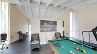 This single-story space is designed as a dedicated recreational and entertainment area, combining casual comfort with functional design. The room's most notable architectural feature is its vaulted ceiling with white-painted exposed wood beams, which adds visual height and an open, airy character to the interior. Dark hardwood flooring grounds the space and provides a rich contrast to the bright, neutral walls and ceiling.

At the center of the room, a full-sized billiard table with a green felt surface serves as the focal point, accompanied by cues and billiard accessories ready for use. Woven rattan lounge chairs are positioned around the room, offering comfortable seating for guests. A striking triptych wall art piece depicting a dramatic ocean seascape adds a bold decorative element to the main wall.

Natural light enters through tall windows and a glass door on the right side of the room, contributing to the bright and welcoming atmosphere. An additional doorway on the left side hints at further connected spaces. The overall ambiance is relaxed yet purposeful, making this an ideal setting for social gatherings and recreational activities.