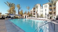 This outdoor amenity space is defined by its resort-style design and Mediterranean-inspired architecture. The centerpiece is a generously sized swimming pool with crystal-clear blue water, framed by a wide, paved sundeck lined with rows of lounge chairs — ideal for relaxing under open skies. Towering palm trees and mature deciduous trees are thoughtfully positioned throughout the area, providing both shade and a lush, landscaped atmosphere.

The surrounding building features a classic stucco exterior in warm neutral tones, with arched windows, tiered balconies, and decorative architectural detailing that lend the space an upscale, cohesive aesthetic. A poolside cabana structure adds a functional and stylistic focal point to one end of the deck, while additional yellow market umbrellas offer shaded retreats around the perimeter.

Adjacent to the pool area is an intimate courtyard patio finished with herringbone-patterned brick pavers. This seating area is furnished with weather-resistant tables and chairs beneath large yellow umbrellas, surrounded by potted planters and climbing greenery along the iron fencing. Drought-tolerant native plantings and decorative landscaping complement the stucco archways, creating a welcoming and well-maintained outdoor retreat throughout the space.