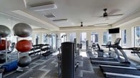 This single-floor fitness center is designed to provide a comprehensive workout experience in a bright, well-appointed environment. Large windows line the walls, flooding the space with natural light and creating an open, airy atmosphere. Ceiling fans are strategically placed throughout to maintain comfortable air circulation during workouts.

The facility is generously stocked with a full array of strength and cardio equipment, including cable machines, weight benches, treadmills, and stationary bikes. A dedicated free weights section features multiple dumbbell racks organized with a wide range of weight options to accommodate all fitness levels. Floor-to-ceiling mirrors are mounted along one wall, allowing users to monitor their form while exercising and visually expanding the sense of space.

The flooring consists of patterned carpet tiles, offering both comfort underfoot and durability suited to a high-traffic fitness environment. The ceiling features crown molding details and recessed HVAC vents, contributing to both climate control and a polished aesthetic. The overall ambiance is clean, modern, and functional, making this fitness center a well-rounded facility suitable for a variety of training routines.