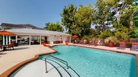Located at 505 Vorils Ln in Wilmington, NC, this outdoor amenity area centers around a generously sized freeform swimming pool with crystal-clear blue water. The pool is framed by a broad concrete sun deck outfitted with vibrant orange lounge chairs and chaise lounges, providing ample space for sunbathing and relaxation. A covered pavilion anchors one end of the pool area, featuring a striking wood-planked vaulted ceiling, stone accent wall, wall-mounted television, and comfortable wicker seating with coordinating orange cushions — creating an inviting shaded retreat that overlooks the pool. The poolhouse structure, finished in white vertical board siding with a standing-seam metal roof, includes what appear to be restroom and utility facilities accessible from the deck. The entire area is enclosed by a black iron fence, ensuring privacy and security, while mature trees lining the perimeter provide a natural, park-like backdrop. Decorative potted plants and red umbrella-shaded seating areas add color and character throughout the deck. An accessibility lift is also present at the pool's edge. The space benefits from abundant natural light and open sky views, making it a well-appointed outdoor recreation destination well-suited for community or resort use.