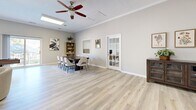 Located at 1300 Gran Crique Dr in Roswell, GA, this well-appointed single-floor space offers a thoughtfully designed open layout that serves multiple functional purposes. Light oak-style luxury vinyl plank flooring runs throughout, complemented by neutral warm-gray walls and crisp white crown molding and baseboards that lend the interior a clean, polished aesthetic.

The living area serves as the focal point of the space, anchored by a striking stacked-stone fireplace with a classic white mantel — an inviting architectural centerpiece. A pair of tufted emerald-green velvet sofas and a round coffee table create an elegant yet comfortable seating arrangement in front of the hearth.

Adjacent to the living area is a dedicated workstation zone, featuring dual computer setups along a wall-mounted desk, ideal for productivity or administrative tasks. A tall potted plant adds a touch of greenery and warmth to this functional corner.

The dining area accommodates a large dark wood conference-style table surrounded by upholstered modern chairs, making it well-suited for meetings or group gatherings. A framed mirror and botanical wall art enhance the room's character. A glass-paned French door connects to an additional room beyond.

Flush-mounted ceiling light panels and a ceiling fan ensure the space is well-lit and comfortable, contributing to an overall atmosphere that is bright, modern, and welcoming.