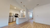 This single-story residence is designed with a modern, open-concept layout that promotes a seamless flow between living spaces. The living room serves as a central gathering area, highlighted by soaring vaulted ceilings with recessed lighting and a tile-surround fireplace as a focal point. Light wood-look flooring runs throughout the main living areas, enhancing the bright, airy atmosphere created by multiple windows fitted with blinds.

The kitchen is well-appointed with white shaker-style cabinetry, light countertops, and a full suite of stainless steel appliances, including a refrigerator, electric range, and over-the-range microwave. A peninsula with pendant lighting overhead provides additional workspace and casual seating, connecting the kitchen to the living area.

Bedrooms feature plush carpeting and windows that bring in natural light and exterior views. The bathroom includes a single-sink vanity with ample cabinet storage, a framed mirror, and a walk-in glass-enclosed shower with wood-look flooring throughout. A dedicated laundry room houses a full-size washer and dryer with overhead cabinetry for added storage.