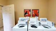 This single-floor laundry facility is designed to provide a practical and comfortable experience for its users. The room is finished with warm, golden-tan painted walls that create an inviting atmosphere, distinguishing it from a typical utilitarian laundry space. Multiple top-loading, coin-operated washing machines are neatly arranged in rows, each equipped with clearly posted instructions for straightforward operation. The machines appear to be well-maintained, reflecting the overall cleanliness of the facility. Adding a notable decorative touch, two framed pieces of artwork featuring bold red backgrounds and striking black silhouette figures are displayed on the wall above the machines, lending the space a degree of character and visual interest uncommon in shared laundry facilities. The layout is organized to allow easy access to each machine, and the overall design balances functionality with a welcoming aesthetic. The space presents itself as a thoughtfully maintained communal amenity that prioritizes both practicality and a pleasant environment for daily use.